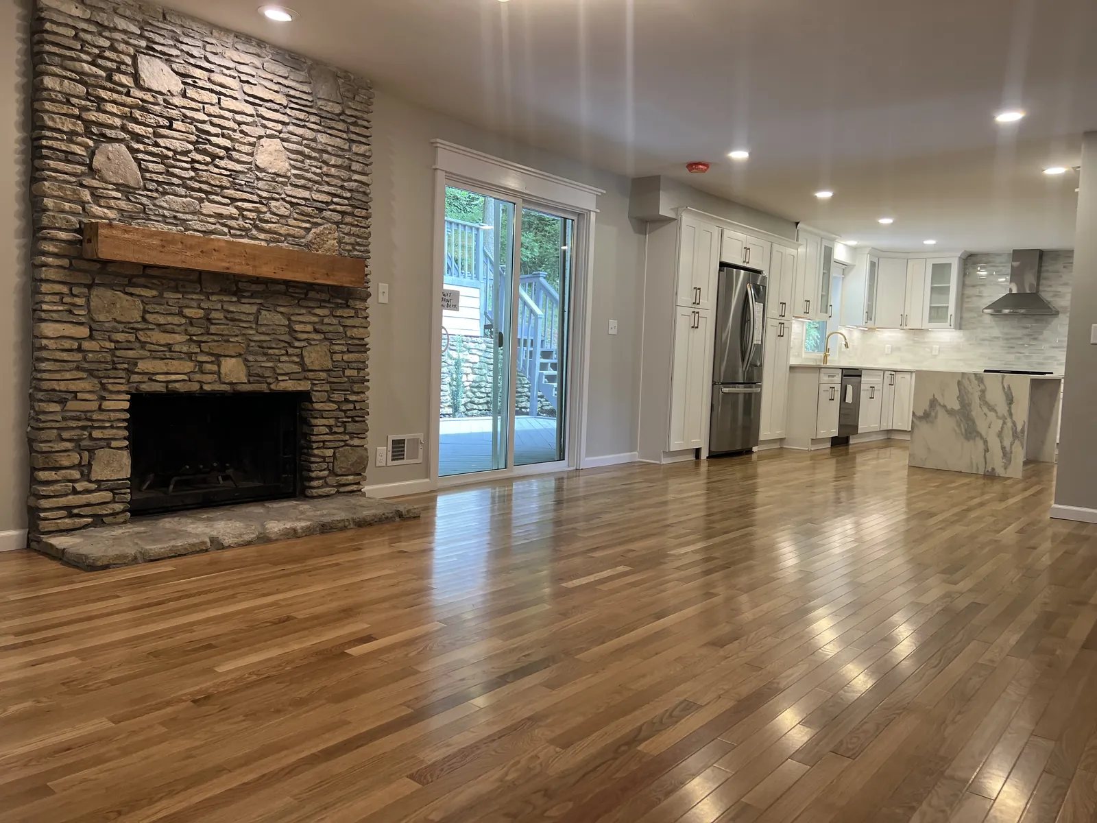 Open living room with red oak flooring, a stacked stone fireplace at left and the kitchen visible beyond