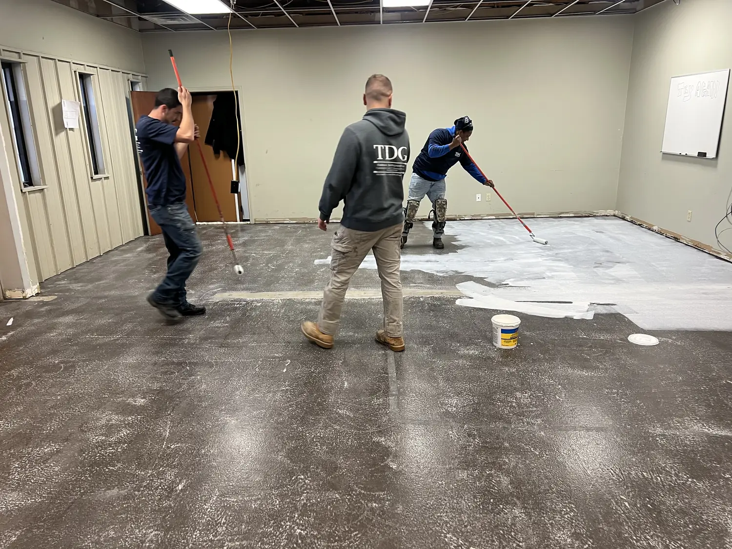 Three-man crew in Trademark Development Group shirts beginning to roll primer over the abraded epoxy floor